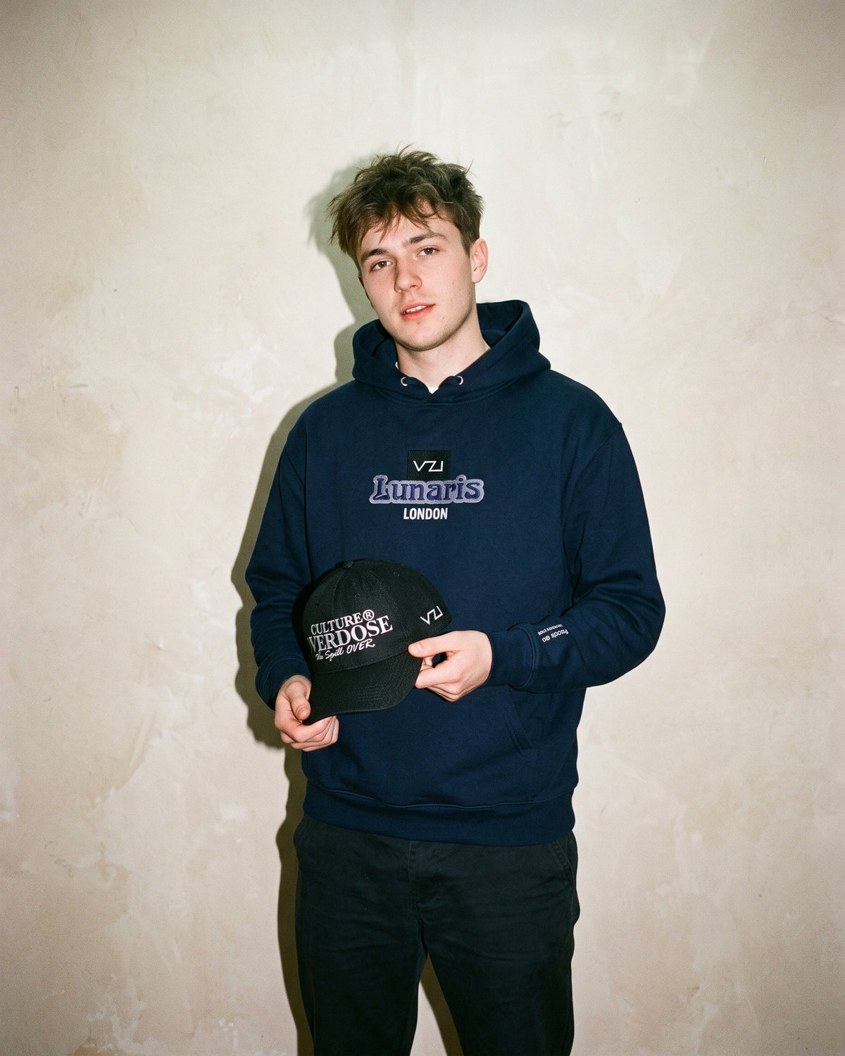 Model in the navy Vazzari Lunaris hoodie with VZI / Lunaris / LONDON chest logo, holding a black Culture Overdose cap against a warm plaster wall
