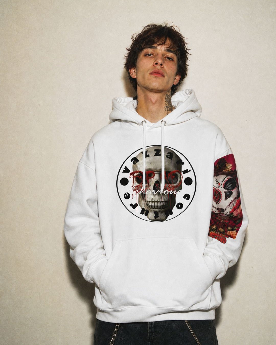 Model in the white Vazzari Deadly Behaviour hoodie with the skull seal print and sugar-skull sleeve print, in a beige studio