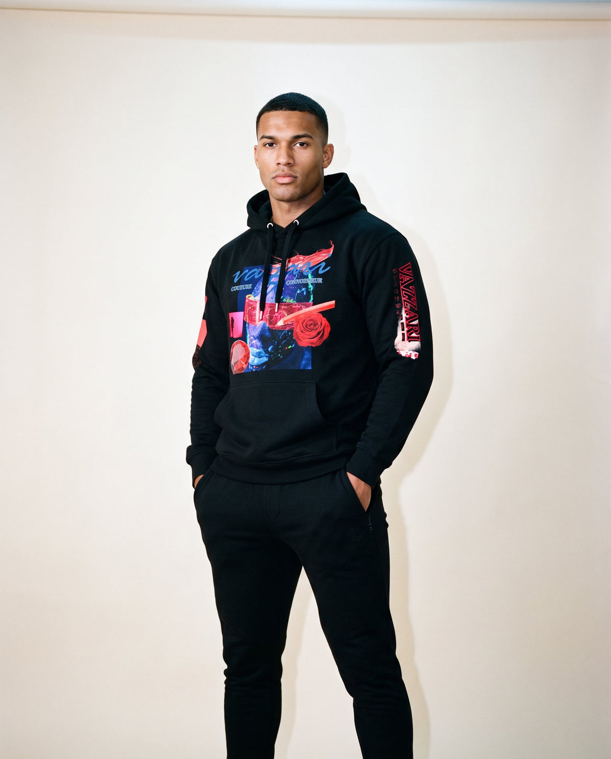 Model in the black Vazzari Transmutation Matrix hoodie with blue-face collage graphic and red VAZZARI sleeve print, in a beige studio
